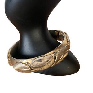 May include: A silver-toned hinged bangle bracelet with a detailed feather design. The bracelet is displayed on a black mannequin hand, showcasing the intricate texture and style of the jewelry.