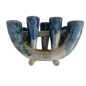 Könnte beinhalten: Ein Kerzenhalter aus Keramik mit einem einzigartigen Design. Der Halter hat eine gebogene, offene Form mit mehreren zylindrischen Öffnungen für Kerzen. Das Stück ist in Blau- und Weißtönen glasiert und hat eine strukturierte Oberfläche. Es steht auf drei kleinen Füßen.