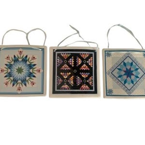 May include: Three square decorative ornaments with quilt-inspired designs. The ornaments feature floral and geometric patterns in shades of blue, pink, and green, with white borders and hanging loops. The designs are reminiscent of traditional quilt blocks.