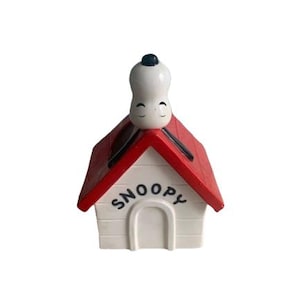 May include: A Snoopy doghouse figurine with a red roof and white walls. The word "SNOOPY" is arched above the door. Snoopy is perched on top of the roof, with his eyes closed. A charming collectible.