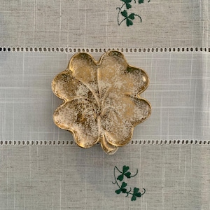 Puede incluir: Un plato dorado y blanco en forma de trébol de cuatro hojas descansa sobre un camino de mesa de lino blanco y gris. El plato tiene una superficie texturizada con detalles dorados. El camino de mesa tiene tréboles verdes bordados y costuras decorativas.
