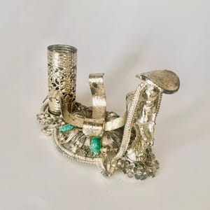 May include: Silver-toned decorative stand with a filigree design and green gemstone accents. The stand has a cylindrical holder and a curved base.
