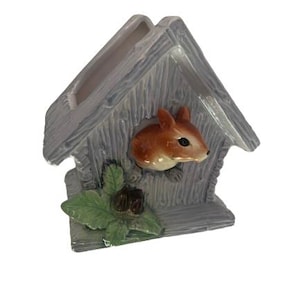May include: A ceramic birdhouse-shaped planter with a gray, weathered wood appearance. A brown squirrel peeks out from a round opening. Green leaves and a brown nut adorn the base. The planter is designed for small plants.
