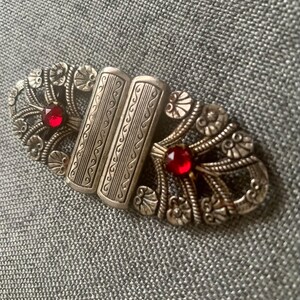 May include: Vintage silver-toned metal clasp with ornate floral and scrollwork design. The clasp features two rectangular center pieces and two red gemstone accents. A decorative accessory.