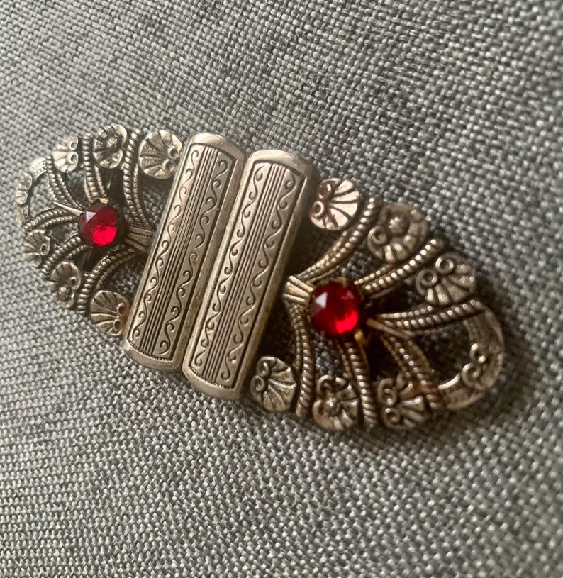 May include: Vintage silver-toned metal clasp with ornate floral and scrollwork design. The clasp features two rectangular center pieces and two red gemstone accents. A decorative accessory.