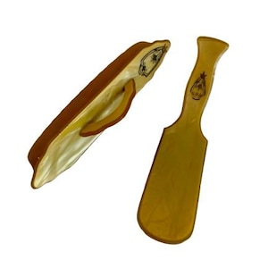 May include: Two vintage shoe horns. One is a curved shoe horn with a handle, and the other is a flat shoe horn. Both are a light yellow color with brown trim and decorative emblems.