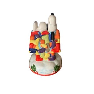 May include: A ceramic Snoopy figurine, wearing a colorful patchwork outfit, stands atop a snowy base with holly accents. The base reads "Christmas 1981". The figurine features a black tie and a white head.