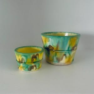 May include: Two ceramic planters with an abstract design in green, yellow and brown. The smaller planter is in front of the larger planter.