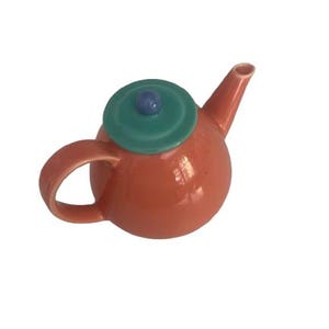 Può includere: Una teiera in ceramica rotonda con un corpo color corallo aranciato lucido, un manico curvo e un beccuccio affusolato. Il coperchio è di un colore verde acqua a contrasto e presenta un piccolo pomello viola, rotondo e liscio, al centro.