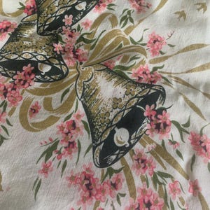 May include: A white fabric with a repeating pattern of gold bells, pink flowers, and green leaves. The bells have a dark green outline and are adorned with gold accents. The flowers are small and clustered, creating a delicate floral design.