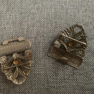 May include: Two antique silver-toned metal clasps with intricate floral and scrollwork designs. Each clasp features a decorative bar and a pin for secure fastening. The clasps have a vintage aesthetic.