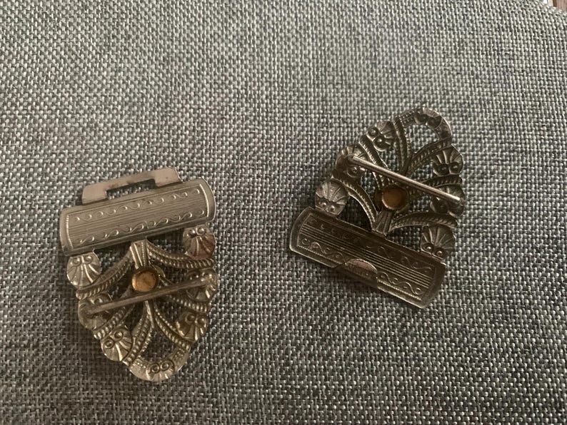 May include: Two antique silver-toned metal clasps with intricate floral and scrollwork designs. Each clasp features a decorative bar and a pin for secure fastening. The clasps have a vintage aesthetic.