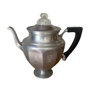 May include: A vintage silver-toned teapot with a clear glass knob on the lid and a black handle. The teapot has an octagonal body with engraved initials. The spout is curved, and the base is a small pedestal.
