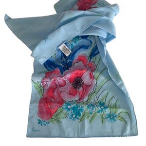 May include: A pale blue silk scarf with a floral pattern. The scarf displays large pink and red flowers with blue accents and green leaves. The scarf is rectangular and appears to be made of a soft, flowing material.