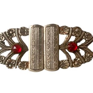 May include: An antique silver-toned buckle with an ornate design. The buckle features two red, faceted gemstones and intricate floral and rope-like detailing. The central clasp has vertical etched lines.
