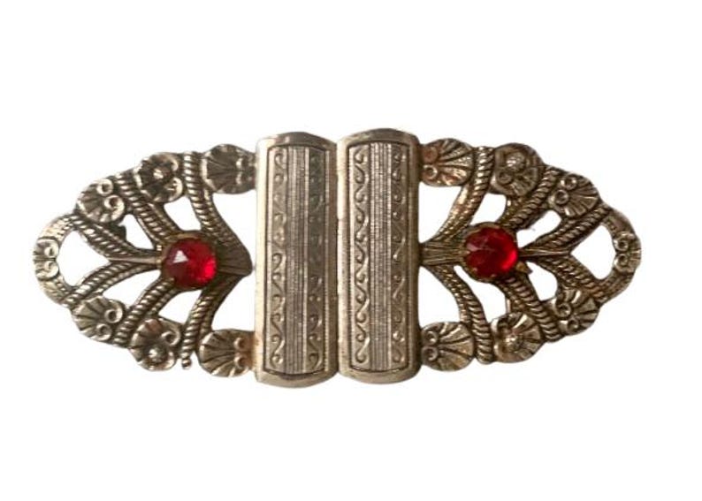 May include: An antique silver-toned buckle with an ornate design. The buckle features two red, faceted gemstones and intricate floral and rope-like detailing. The central clasp has vertical etched lines.