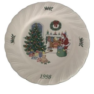 May include: A white decorative plate with a green rim and a swirling edge. The plate features a Christmas scene with a decorated tree, Santa Claus, a fireplace, and the year "1998".