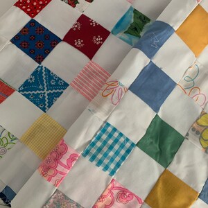 May include: Close-up of a handmade quilt with a patchwork design. The quilt features a variety of colorful fabric squares, including red, blue, pink, and yellow, separated by white fabric strips. The squares have floral, geometric, and gingham patterns.