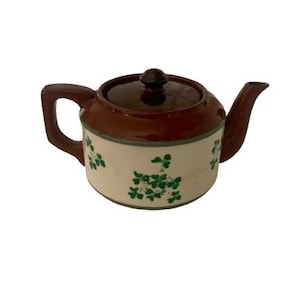 May include: A ceramic teapot with a brown lid, handle, and spout. The body is cream-colored with green shamrock designs. A dark brown knob sits atop the lid. The teapot is designed for brewing tea.