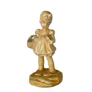 Peut inclure: Une figurine en céramique d'une fille en robe blanche tenant un panier et une fleur. Elle est debout sur une petite base.