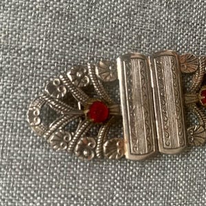 May include: Close-up of an antique silver-toned clasp with ornate floral and scrollwork designs. The clasp features two rectangular bars and is accented with small, round, red gemstones. The clasp is likely for a vintage garment or accessory.
