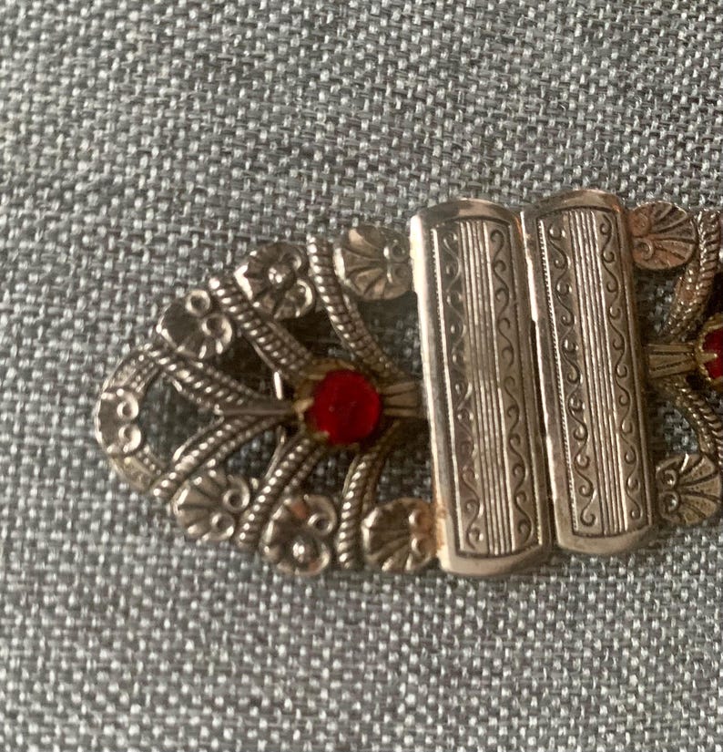 May include: Close-up of an antique silver-toned clasp with ornate floral and scrollwork designs. The clasp features two rectangular bars and is accented with small, round, red gemstones. The clasp is likely for a vintage garment or accessory.