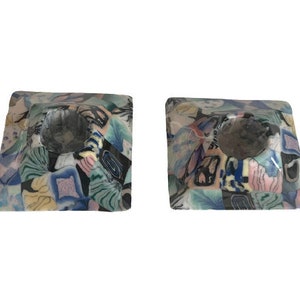 May include: Two square candle holders with a colorful, abstract design. The holders feature a mix of blue, green, pink, and yellow patterns. Each holder has a circular indentation for a candle. The background is white.