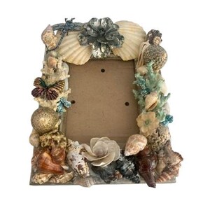 May include: Decorative photo frame adorned with seashells, starfish, and a dragonfly. The frame features a variety of shells in shades of brown, beige, and white, with accents of blue and green. The frame is approximately 8 inches tall and 6 inches wide.