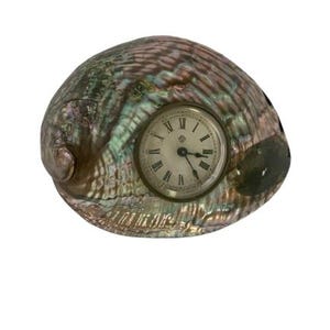 May include: Abalone shell clock with a round face featuring Roman numerals. The shell has iridescent colours of green, pink, and purple. The clock hands are black.