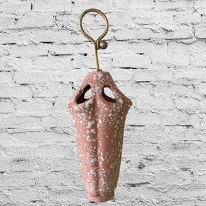 May include: A pink ceramic vase with white speckles and a gold metal hanger. The vase has a unique, geometric shape with cutouts.