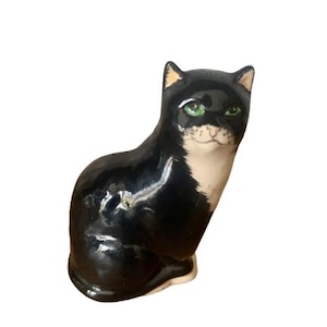 Puede incluir: Figura de cerámica de un gato sentado. El gato es principalmente negro con marcas blancas en el pecho y la cara. Tiene ojos verdes y detalles amarillos en las orejas. La figura es brillante y parece estar pintada a mano.