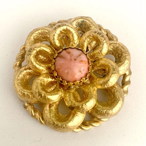 Puede incluir: Un broche de flor de tono dorado con una piedra cabujón rosa en el centro. La flor está hecha de metal retorcido y tiene un borde de cuerda.