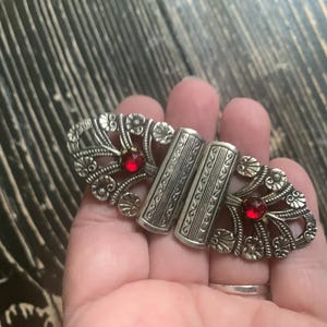 May include: Silver-toned metal clasp with ornate floral and scrollwork designs. The clasp features two rectangular bars and two red gemstone accents. A vintage-style accessory.