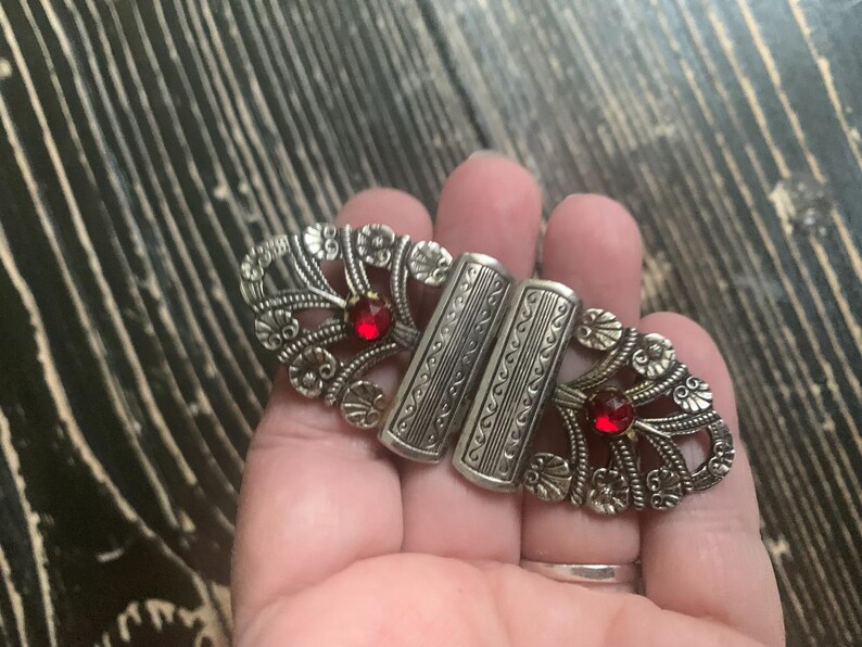 May include: Silver-toned metal clasp with ornate floral and scrollwork designs. The clasp features two rectangular bars and two red gemstone accents. A vintage-style accessory.