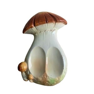 May include: A ceramic mushroom-shaped dish with a brown cap and a white stem. The dish has two oval-shaped compartments and several small mushrooms on the side. The base has green and brown accents.