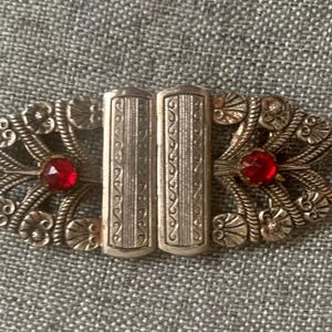 May include: An antique silver-toned brooch with an oval shape. The brooch features two rectangular center pieces and decorative filigree edges. Two red gemstones are set on either side of the center.