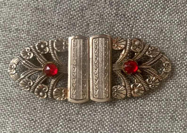May include: An antique silver-toned brooch with an oval shape. The brooch features two rectangular center pieces and decorative filigree edges. Two red gemstones are set on either side of the center.