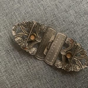 May include: An antique silver-toned belt buckle with an ornate, openwork design. The buckle features two rectangular clasps and decorative oval end pieces with small, round accents. The intricate details suggest a vintage or Art Deco style.