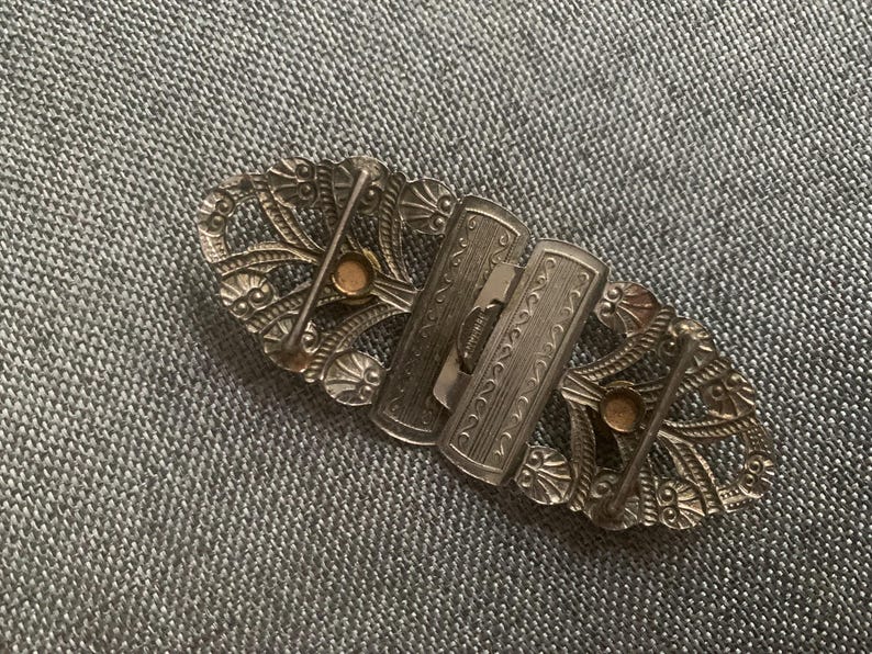 May include: An antique silver-toned belt buckle with an ornate, openwork design. The buckle features two rectangular clasps and decorative oval end pieces with small, round accents. The intricate details suggest a vintage or Art Deco style.