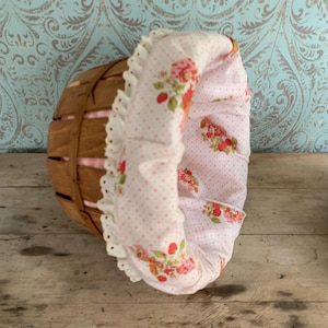 Peut inclure: Un petit panier rond en bois avec une doublure à pois roses et blancs ornée de fraises et de motifs floraux. La doublure a un bord à volants. Le panier est posé sur un fond à motifs de style vintage.