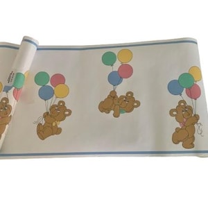 May include: A roll of wallpaper featuring a repeating pattern of brown teddy bears holding colorful balloons. The wallpaper has a light blue border and a white background. The bears are smiling and the balloons are in shades of pink, green, blue, and yellow.