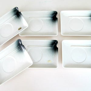 May include: Set of six white ceramic saucers with black and white ombre design. Each saucer has a small indentation for a cup and a raised ridge for a spoon. The saucers are perfect for serving coffee or tea.