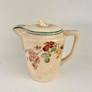 Op de afbeelding: Een crèmekleurige keramische kan met een bloemenmotief. De kan heeft een deksel met een klein vogeltje als knop. Het bloemenmotief heeft roze, gele en groene bloemen.