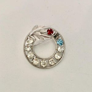 May include: A silver tone brooch with a circle of clear rhinestones and a single red rhinestone, a blue rhinestone, and a gray rhinestone. The brooch has a leaf design on the top.