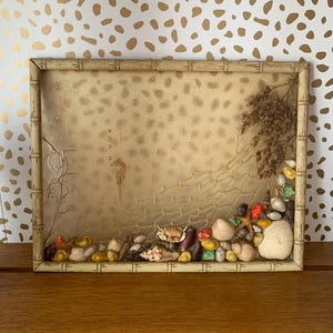 May include: A rectangular decorative frame with a bamboo-style border. The interior features a textured, gold-toned background with a seahorse and other aquatic designs. The frame is adorned with seashells, pebbles, and a starfish.