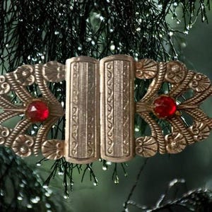 May include: A vintage gold-toned metal buckle with ornate filigree designs and two red gemstone accents. The buckle has a central rectangular clasp. The background is a blurred image of green foliage.