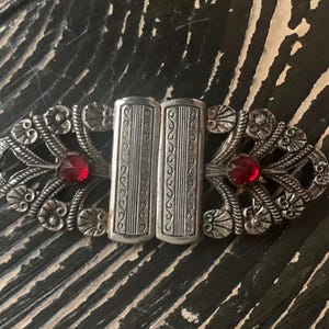 May include: An antique silver-toned clasp with ornate floral and scrollwork designs. Two rectangular center pieces are flanked by decorative elements and bright red, round gemstones. A vintage accessory.