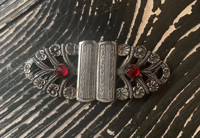 May include: An antique silver-toned clasp with ornate floral and scrollwork designs. Two rectangular center pieces are flanked by decorative elements and bright red, round gemstones. A vintage accessory.
