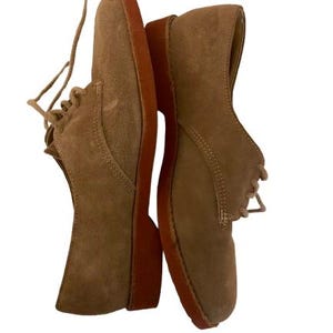 Può includere: Un paio di scarpe Oxford in camoscio color cammello con suole marroni e lacci coordinati. Le scarpe hanno un design classico, adatto per un abbigliamento casual o semi-formale. Il camoscio conferisce loro un aspetto morbido e strutturato.