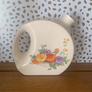 May include: Cream-coloured ceramic pitcher with a large handle and a spout. The pitcher is decorated with a floral arrangement of orange, yellow, and purple flowers. The pitcher is on a wooden surface.